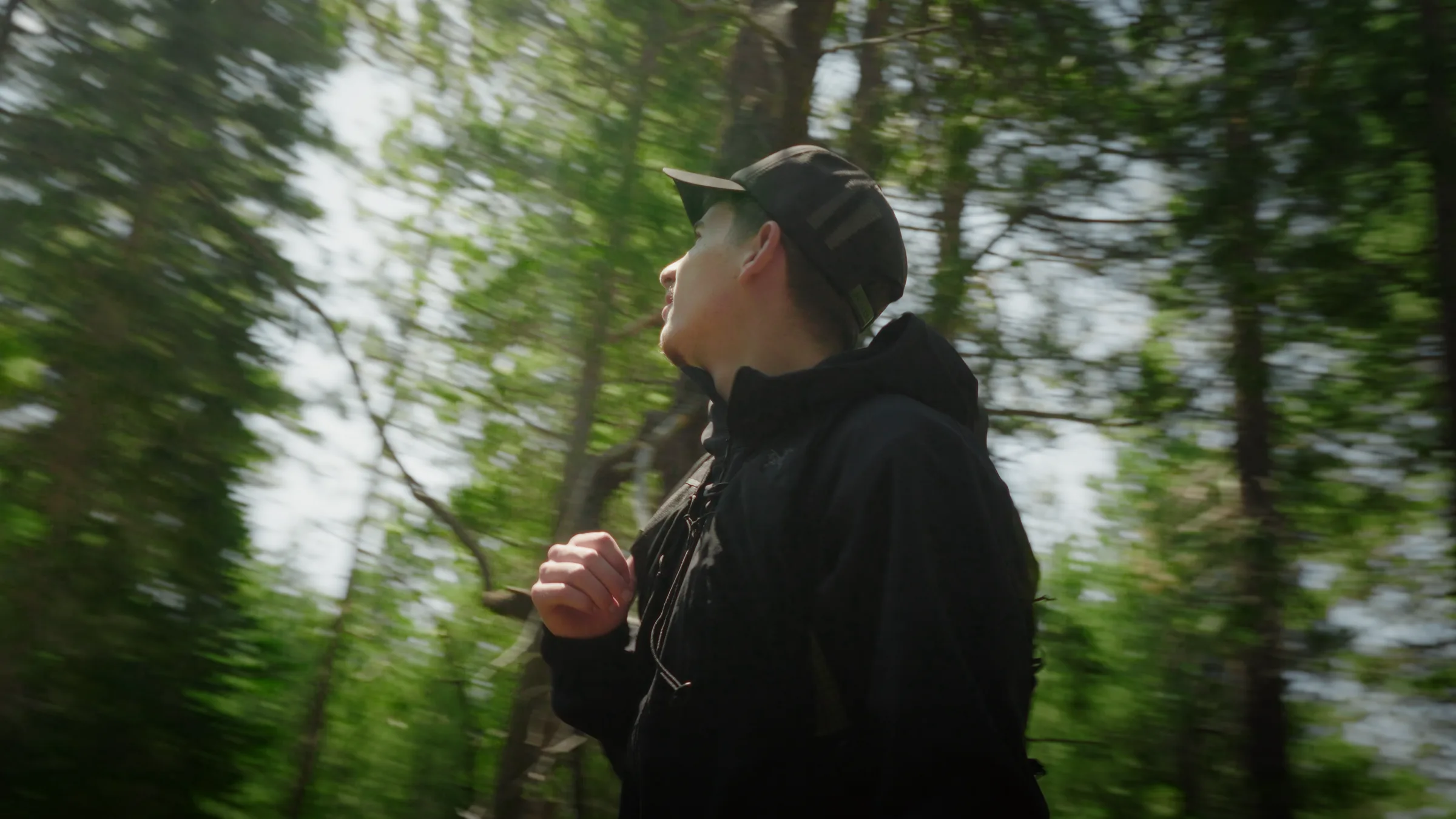 A hiker walks away down a forest trail, motion blur trailing through the pines, an Arc'teryx pack on his back