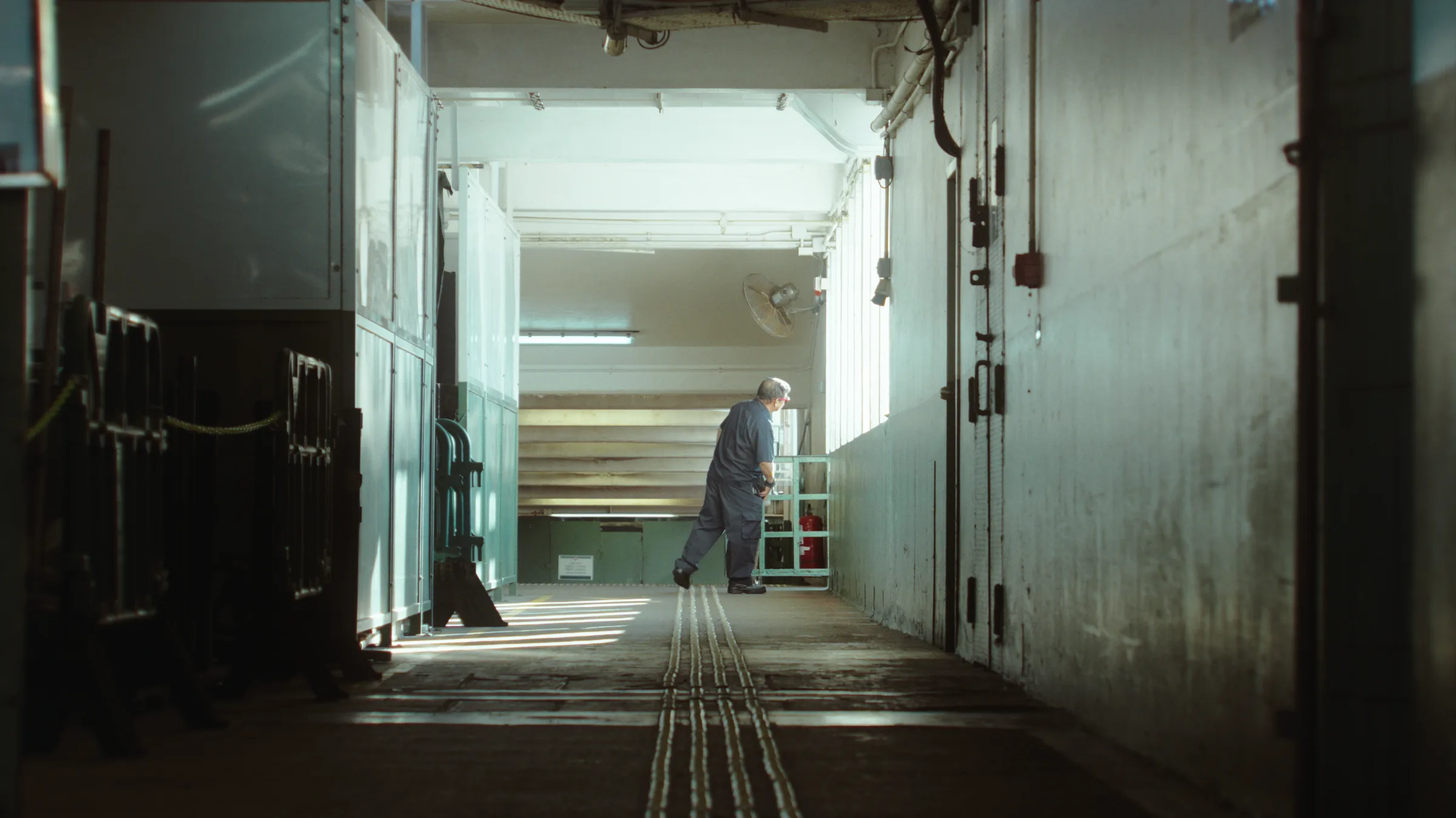 A man walks alone down a sunlit industrial corridor past steel doors and stacked cages