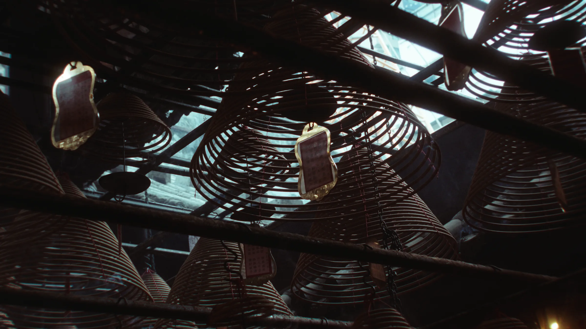 Spiral incense coils and paper prayer tags hang from a temple ceiling, daylight filtering through the roof