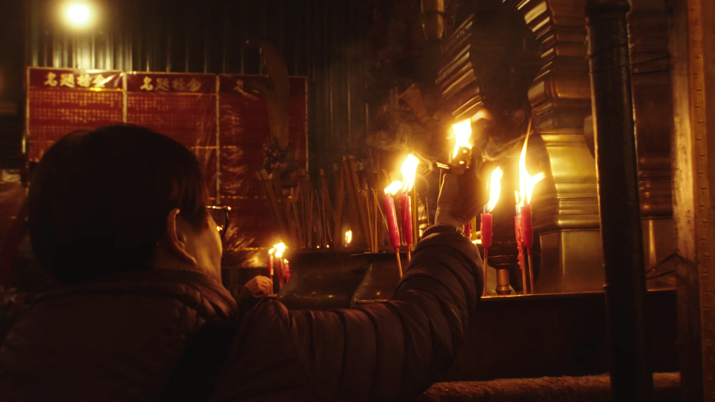 A hand lights a row of temple candles, incense smoke rising through the firelight