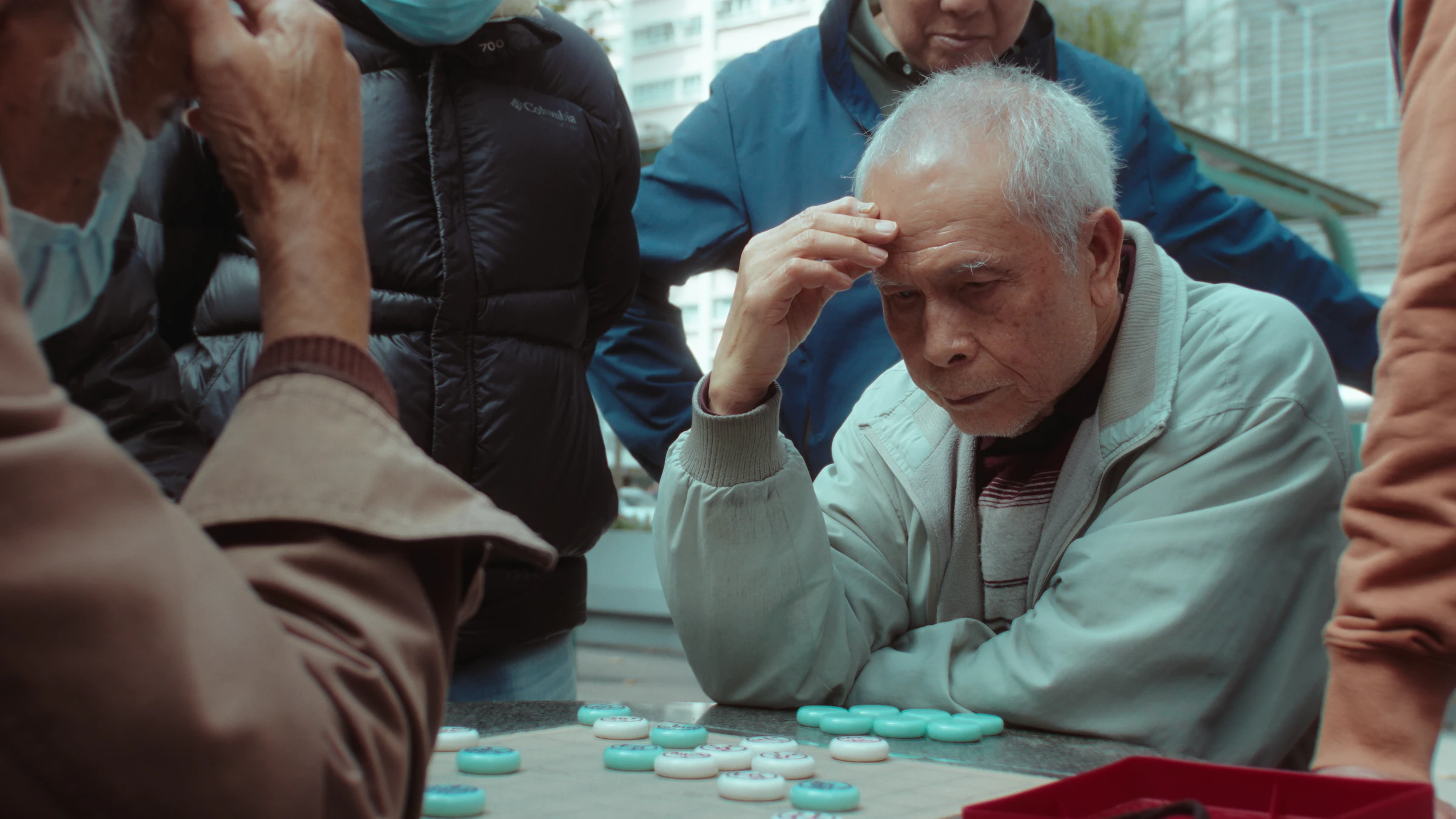 An elderly man rests his head against his hand, deep in thought over a game of Chinese chess at an outdoor table