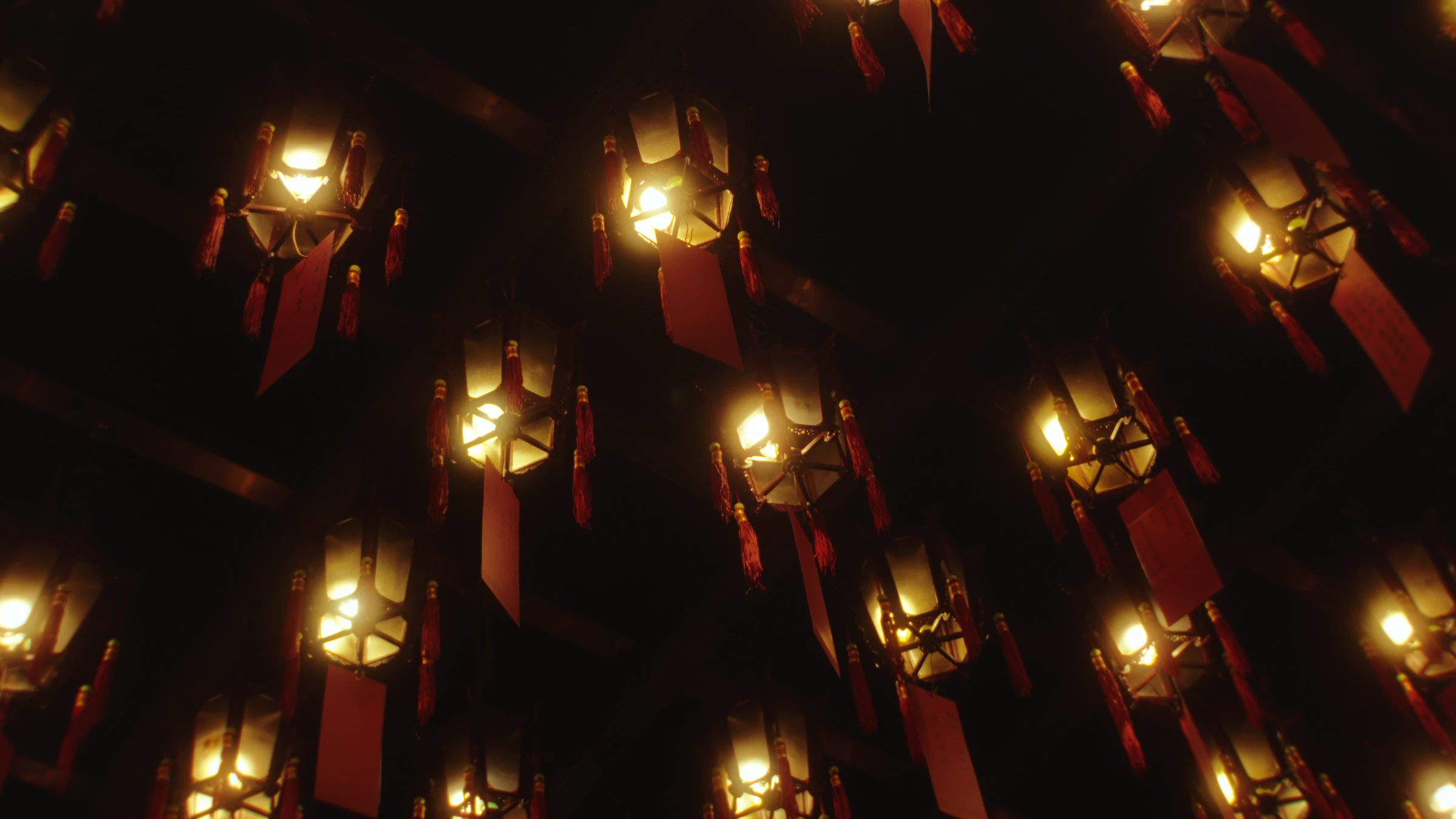 Rows of hanging temple lanterns and red prayer tags glow amid dark ceiling rafters