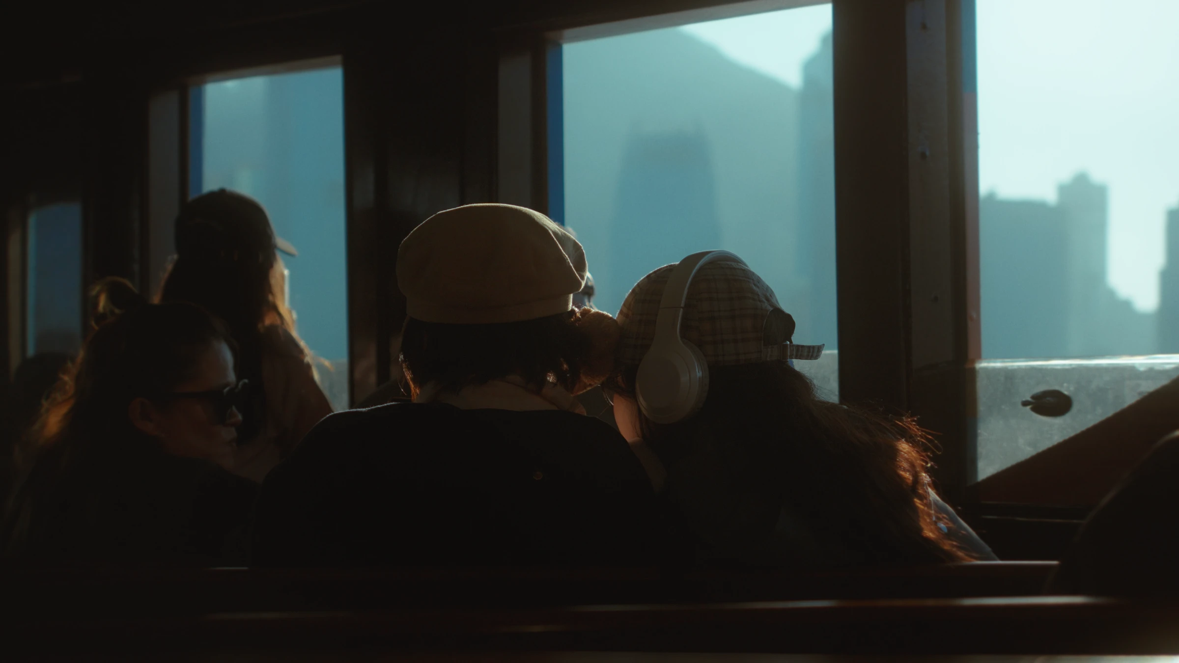 Silhouetted passengers seen from behind inside a ferry cabin, backlit by a hazy Hong Kong skyline through the windows