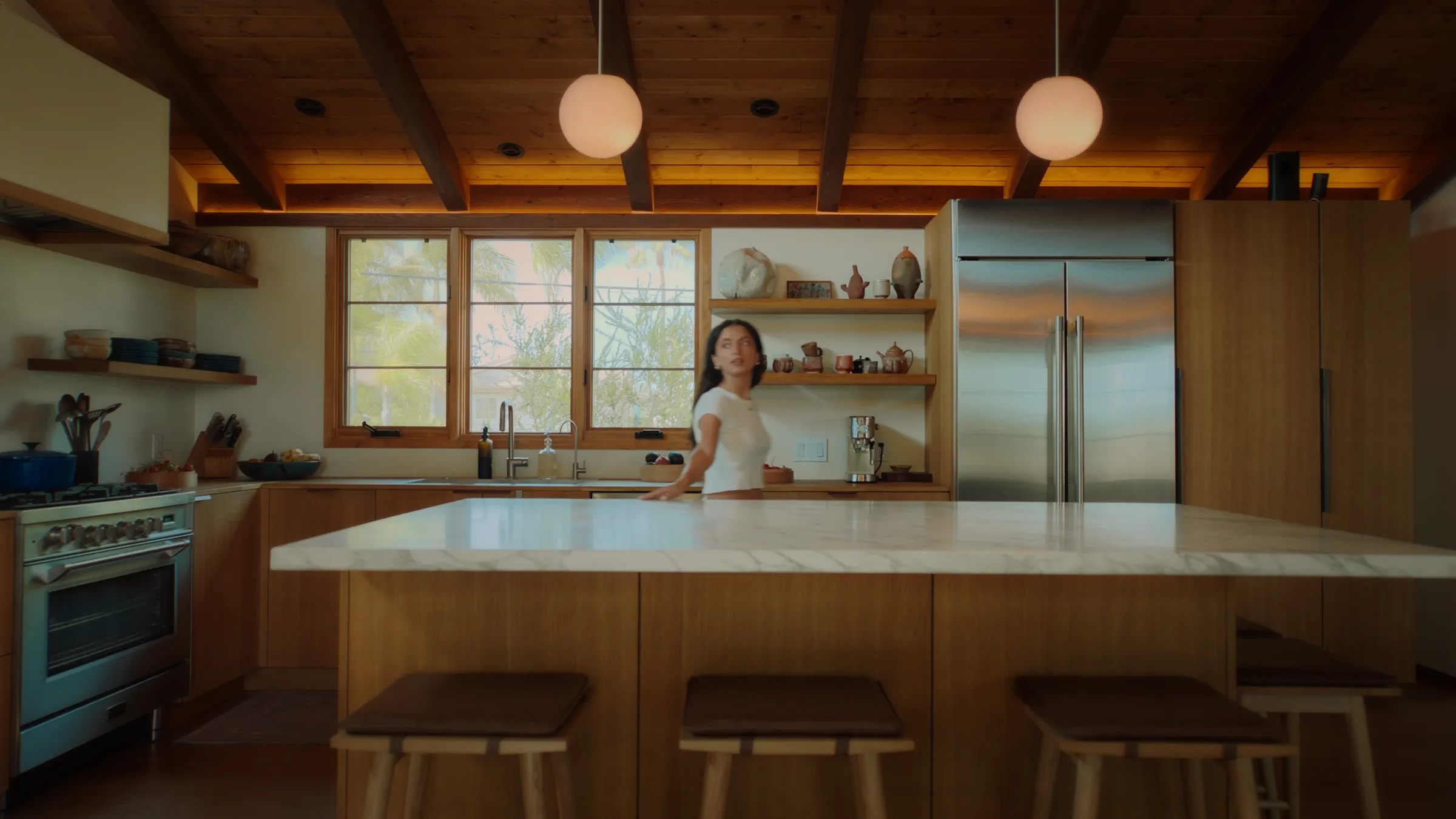 Woman in a white t-shirt caught mid-turn beside a marble kitchen island, wood-beamed ceiling and globe pendant lights behind her