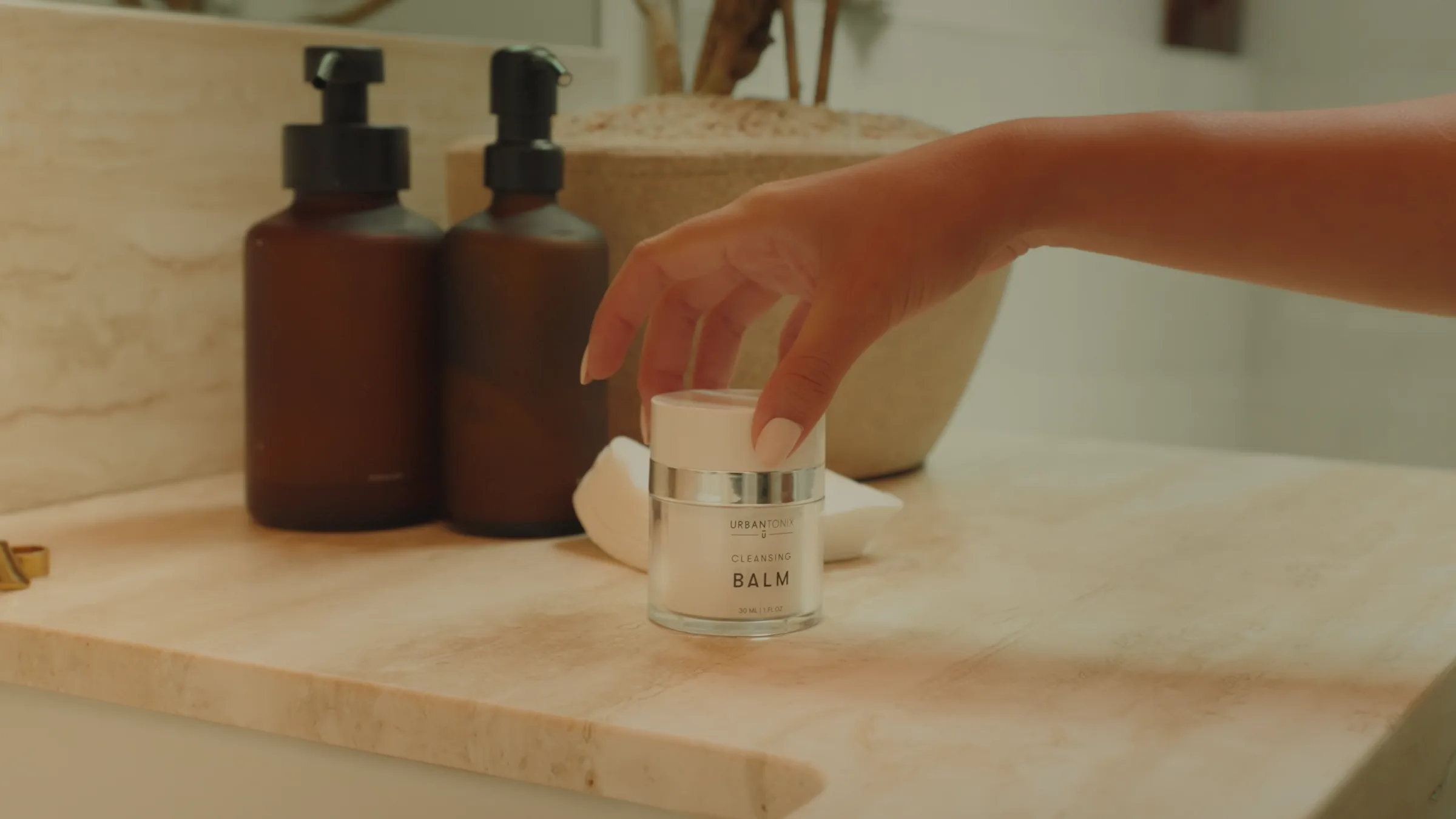 Overhead shot of two hands holding a compact with a dab of amber product, under a brass gooseneck tap over a white sink, gold stacked rings on the counter edge