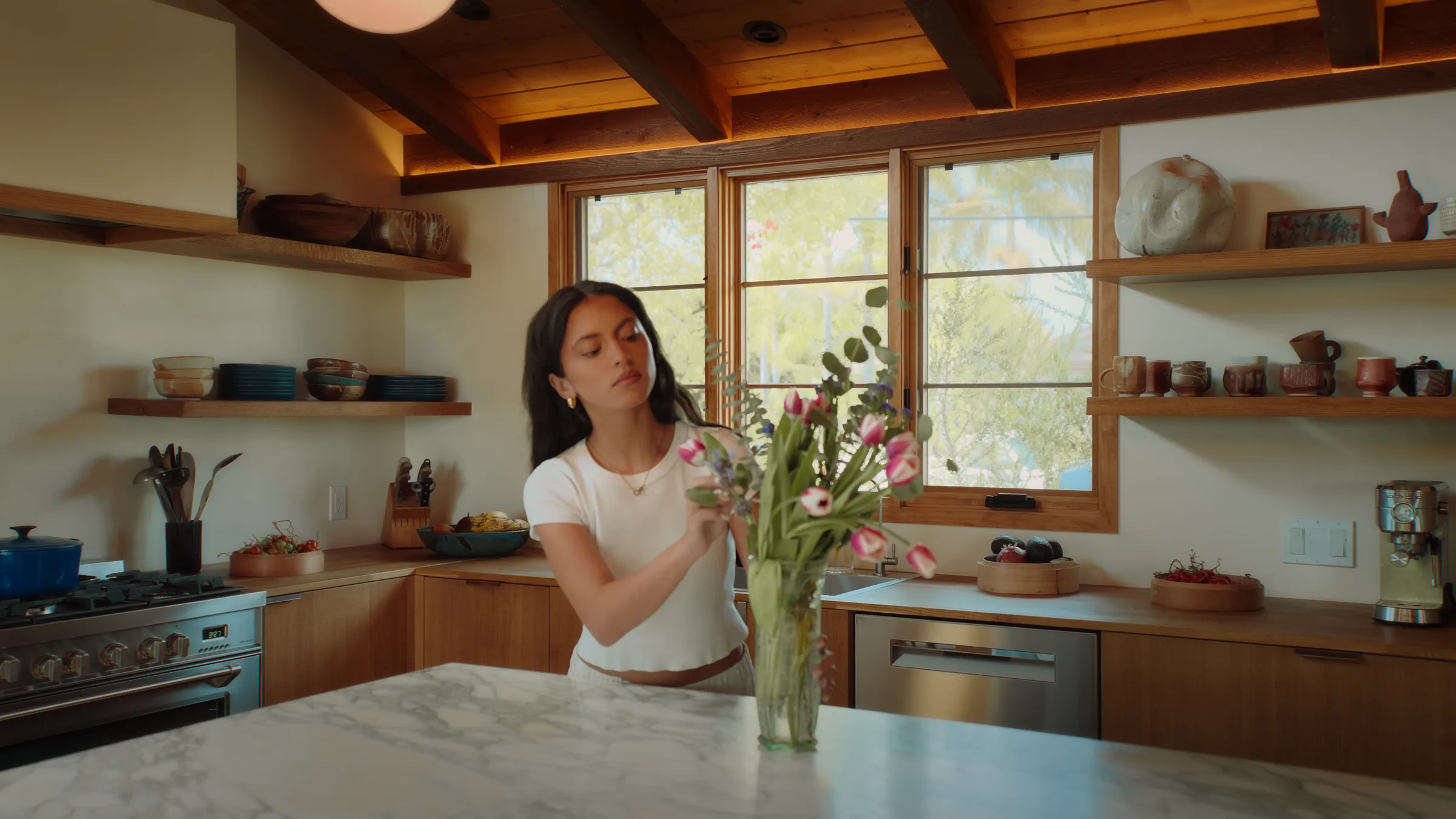 Woman in white top and cream trousers, barefoot, reading a book through open sliding glass doors, potted olive tree and woven lounge chair in the foreground