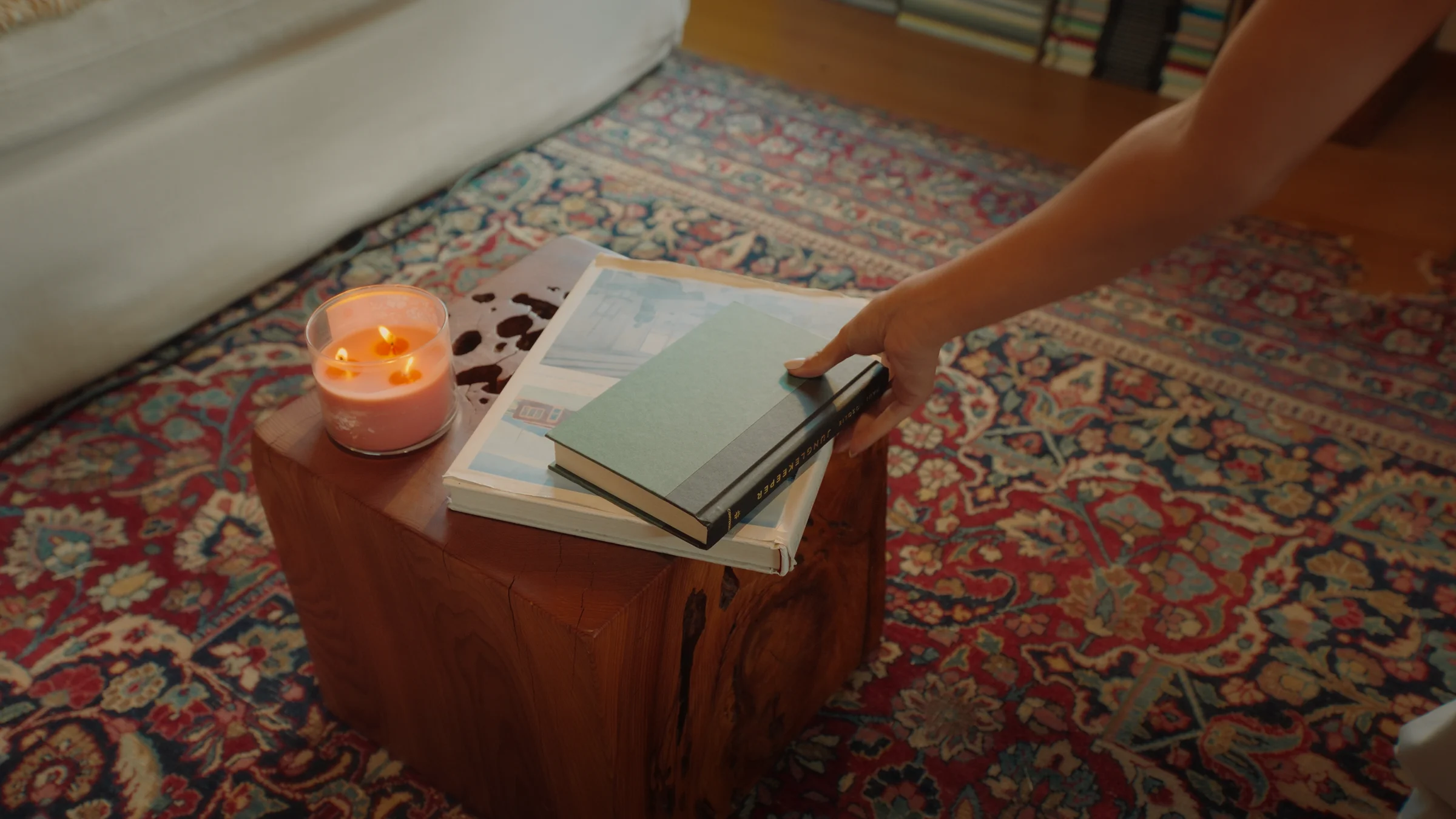 A hand reaches for a stack of books beside a lit candle on a wooden side table, set on a patterned rug beside a cream sofa