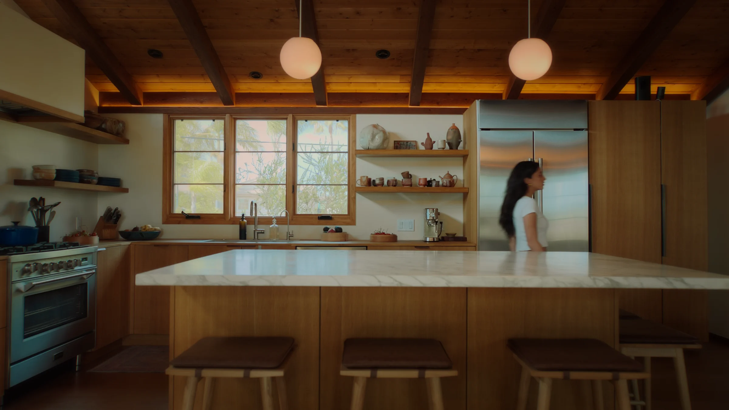 A woman walks through a warm, wood-beamed kitchen at dusk, pendant lights glowing above a marble island